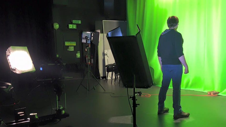A landscape image of a professional Film studio with LGBT Voice Actor Jaden Randall standing next to a green screen with his back to the camera.